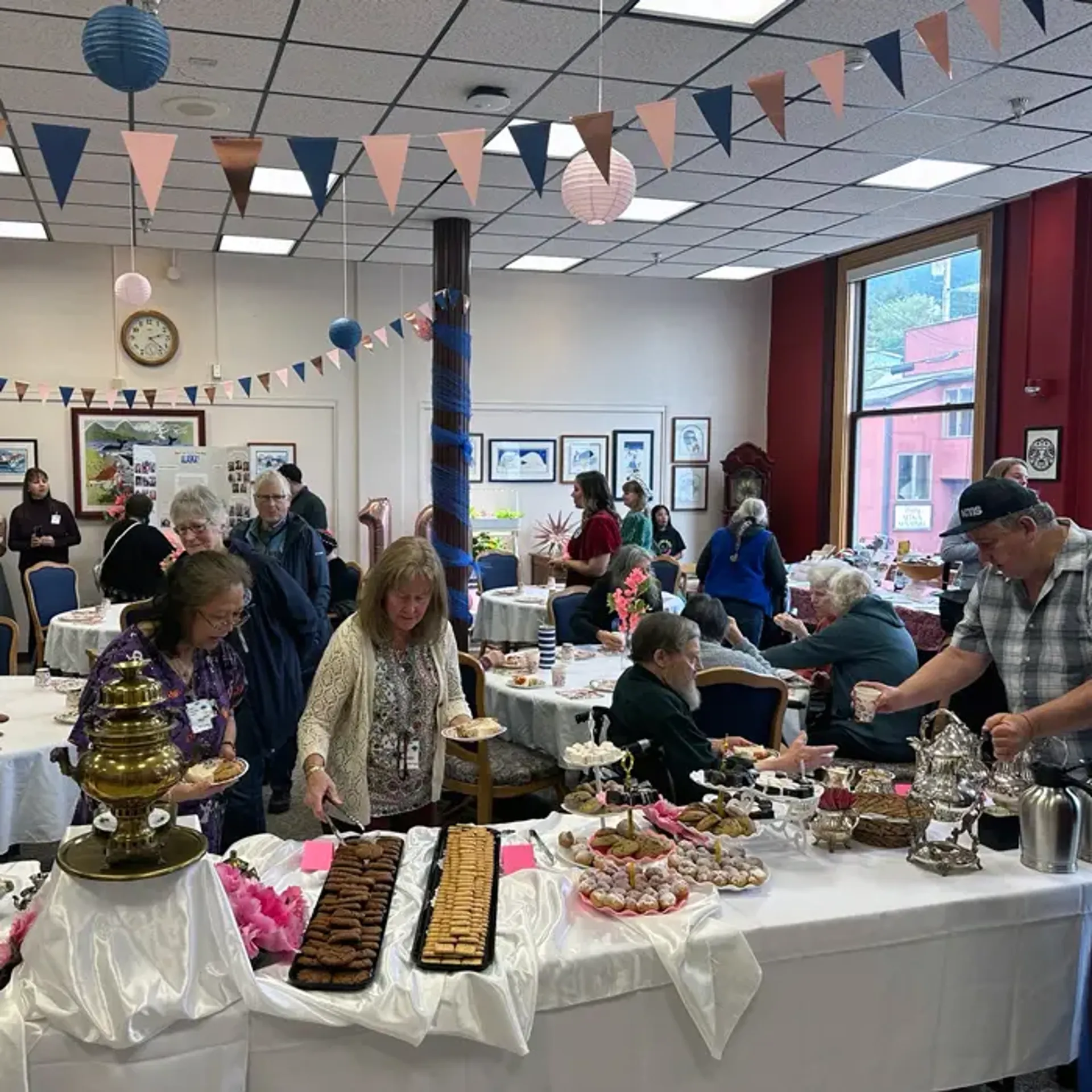 Crowded room, decorated like a party, with people selecting food from a table