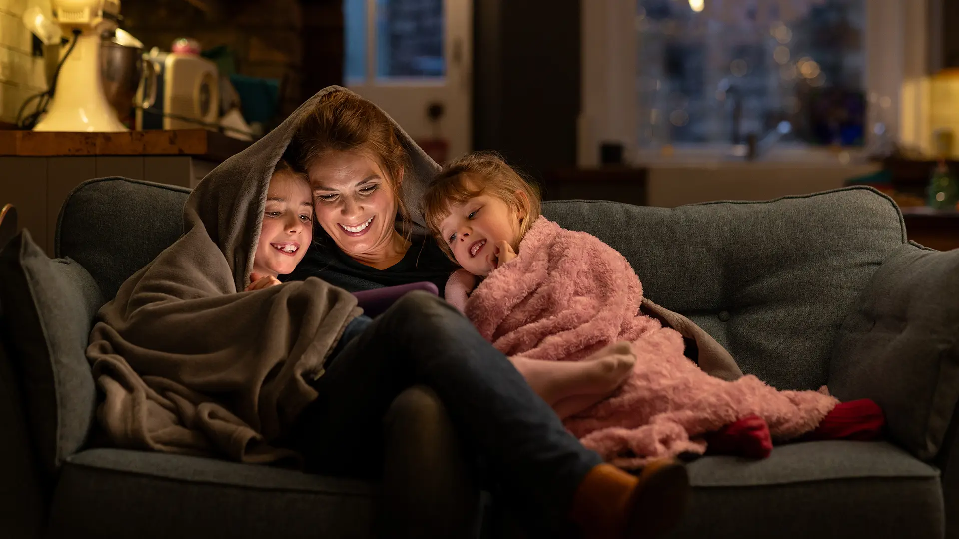 Parent reading to two kids while under a blanket on the couch