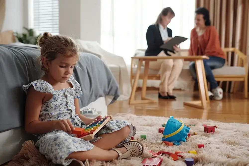 Girl sitting and playing on the floor while her mom talks to a social worker