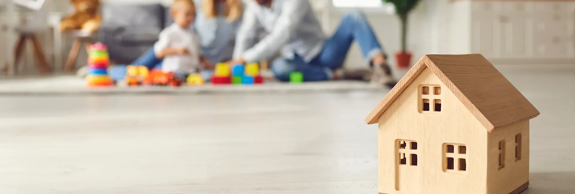 Close up of a small wooden house on the floor, a young family with a child in the background.