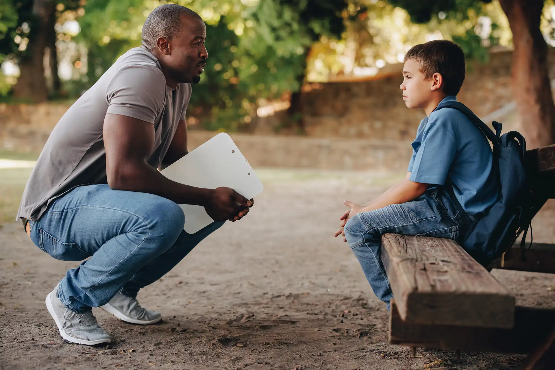 Adult with clipboard crouched down to talk with a sad boy sitting on a bench