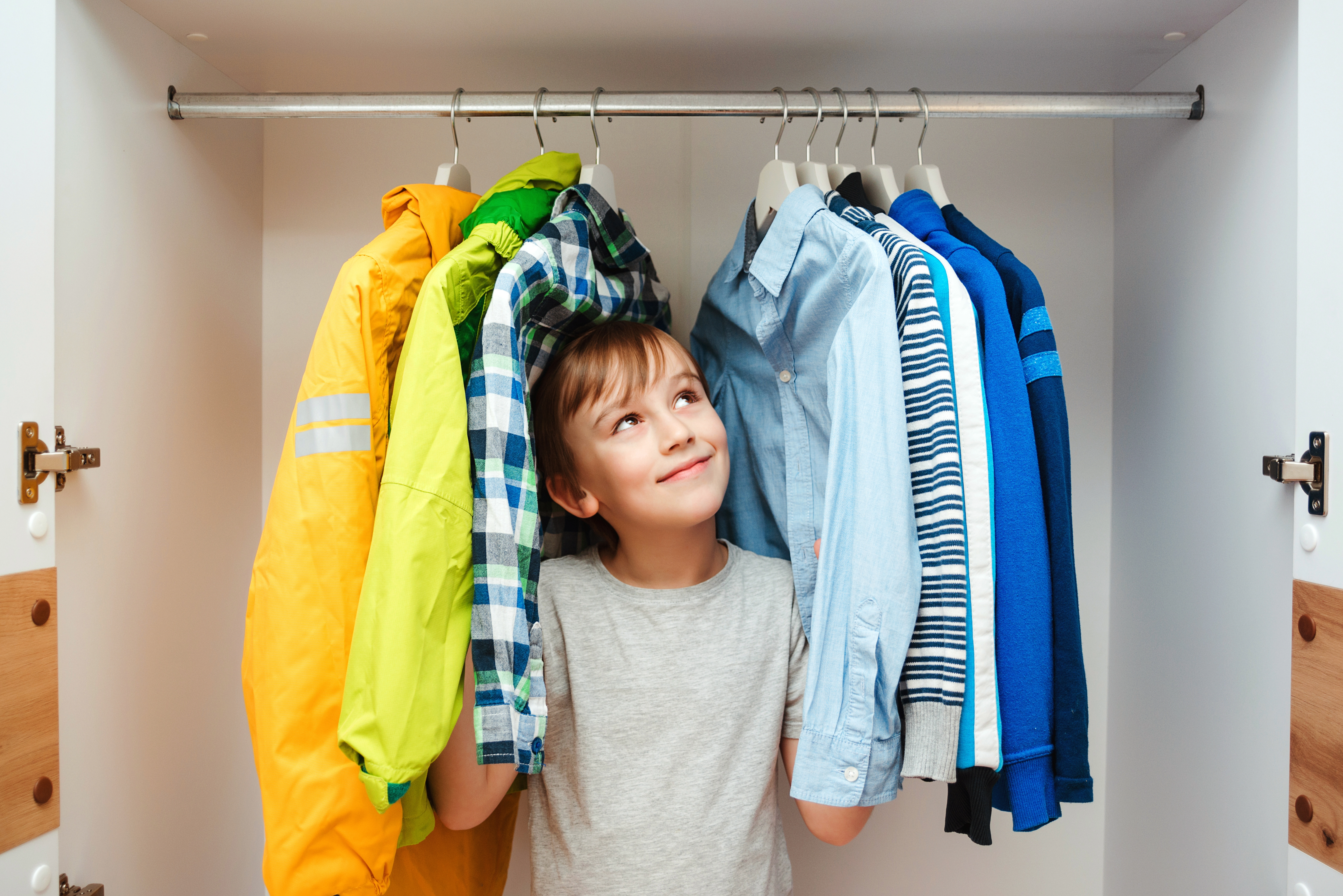 A young boy stands in a closet in between hanging clothes